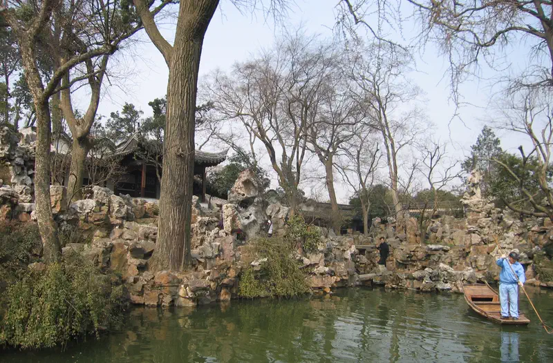 Lingering Garden: rocky courtyard with bare trees beside a pond, and a person walking along a stone path