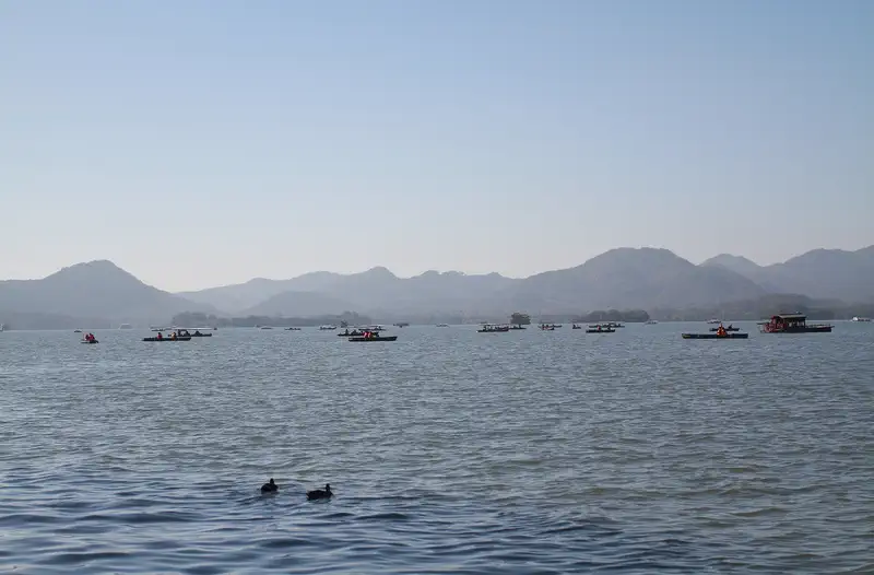 West Lake: calm lake water with small boats and distant hills under a hazy sky