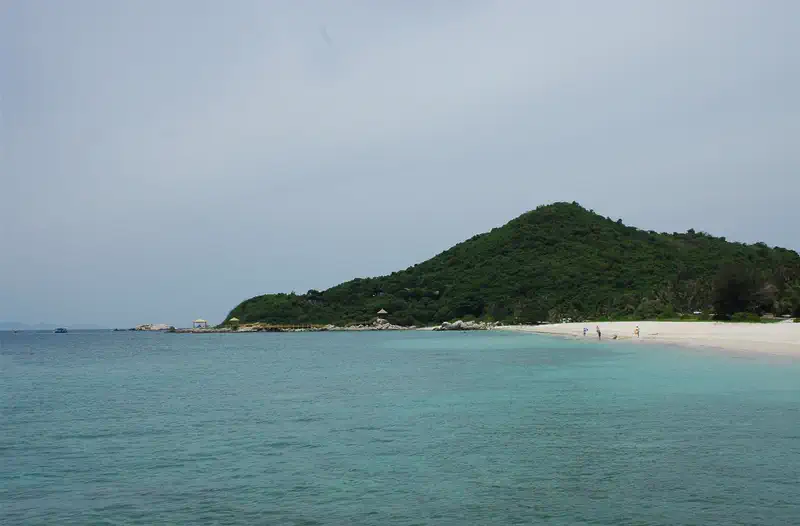 Wuzhizhou Island: turquoise sea water beside a green hill and sandy beach under a hazy sky