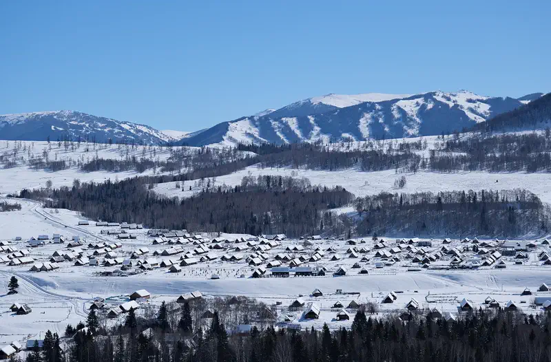 Altay: the log cabins of Hemu village under snow, with forested ridges and white peaks behind