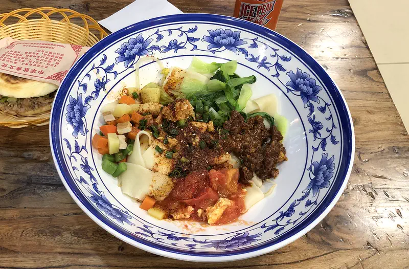 Biangbiang Noodles: wide hand-pulled noodles topped with minced meat, tomato, greens and chilli in a blue-and-white bowl